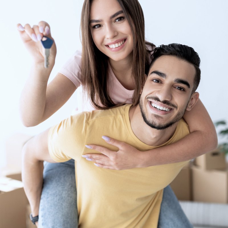 Cheerful Caucasian lady getting piggyback ride from her Arab boyfriend, showing house key indoors. Lovely multinational couple posing in new apartment on moving day